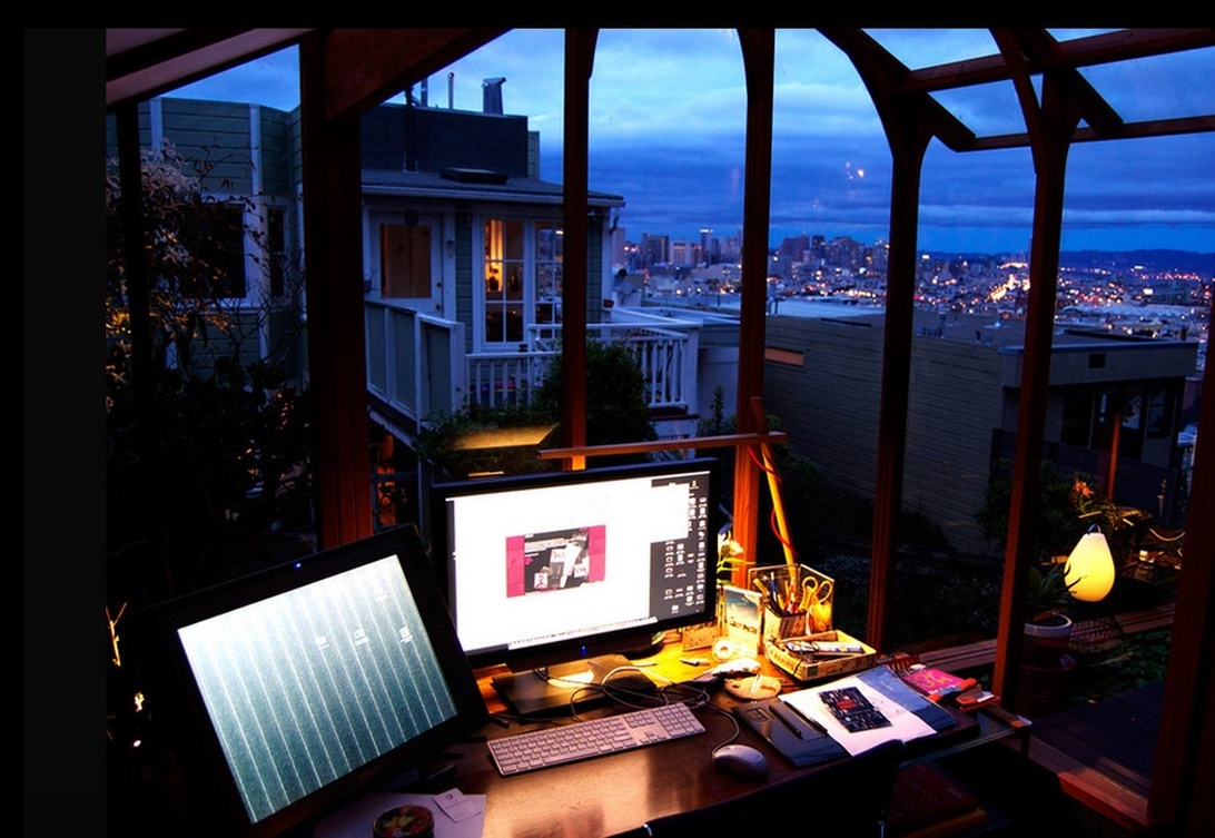 A dramatically lit home office or workspace set in what appears to be a glass-enclosed porch or sunroom with a spectacular nighttime city view. The desk features multiple computer monitors and appears to be set up for technical work. The unique setting with panoramic views represents an innovative approach to workspace design, while the technology suggests possible scientific or technical work.